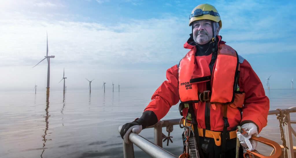Man in a red coat and lifejacket standing on platform next to wind turbines in the sea.